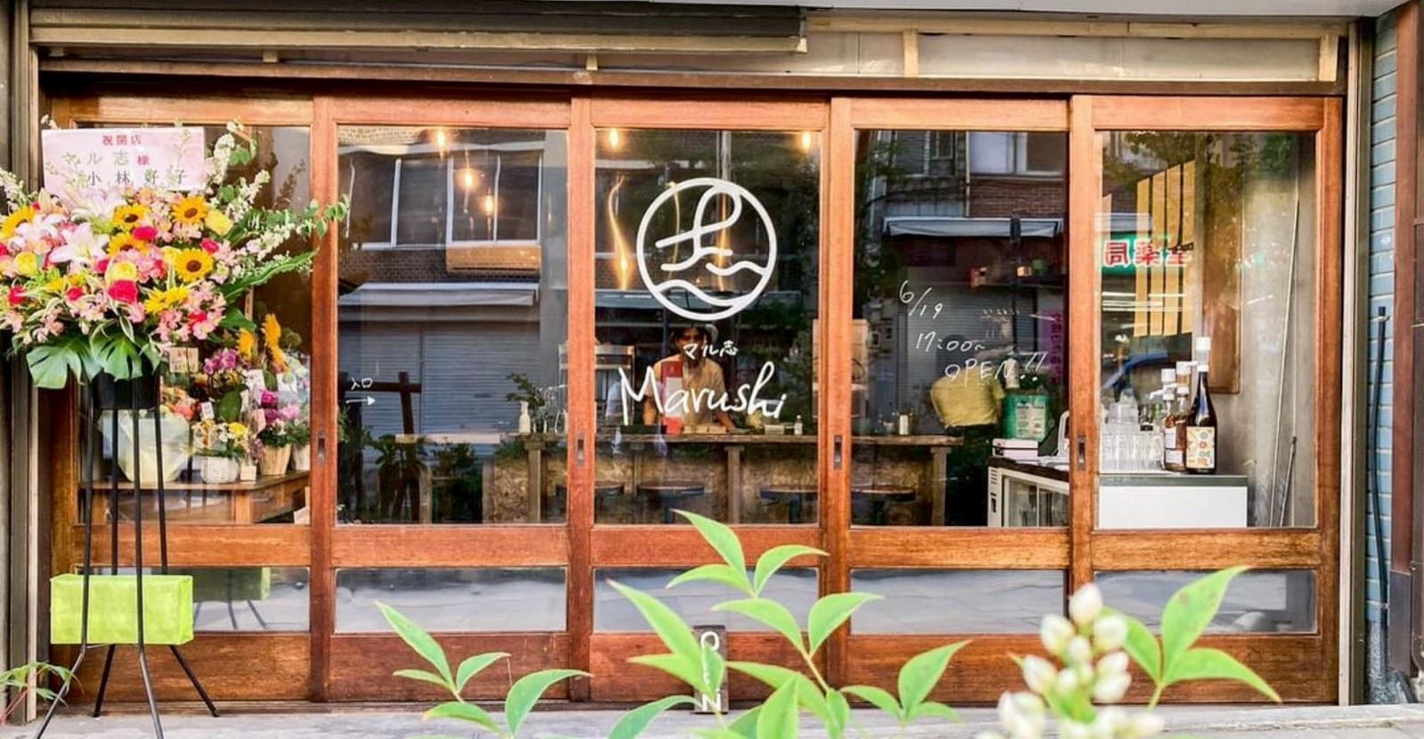 マル志 カレーとおつまみとお酒の外観。ガラス戸に店名ロゴが描かれ、開店祝いの花が並ぶ